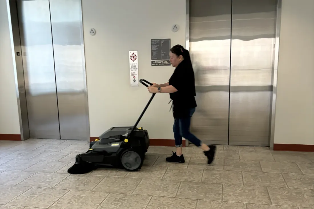 A woman in black pushing a walk-behind floor sweeper across a stone-tile floor past stainless steel elevator doors, EBS staff at work, the same crews that serve Pico Rivera, CA