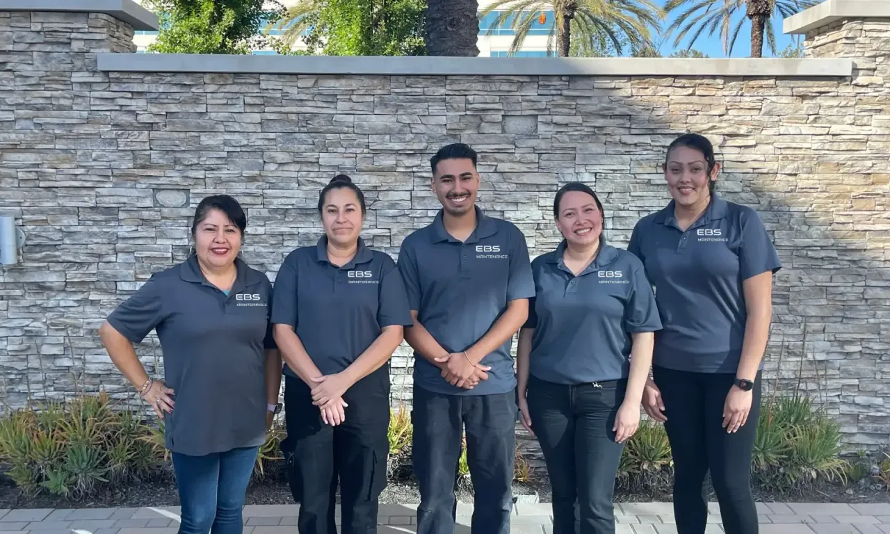 Five team members in matching grey polo shirts with the EBS logo standing in front of a stacked-stone wall, with palm trees behind, EBS staff at work, the same crews that serve Baldwin Park, CA