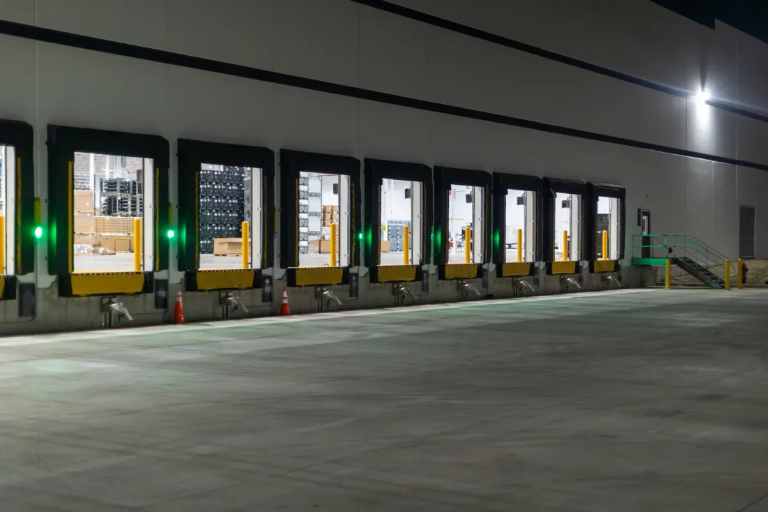 A row of loading dock doors with yellow bumpers and green signal lights along a warehouse wall at night, facing an empty concrete apron