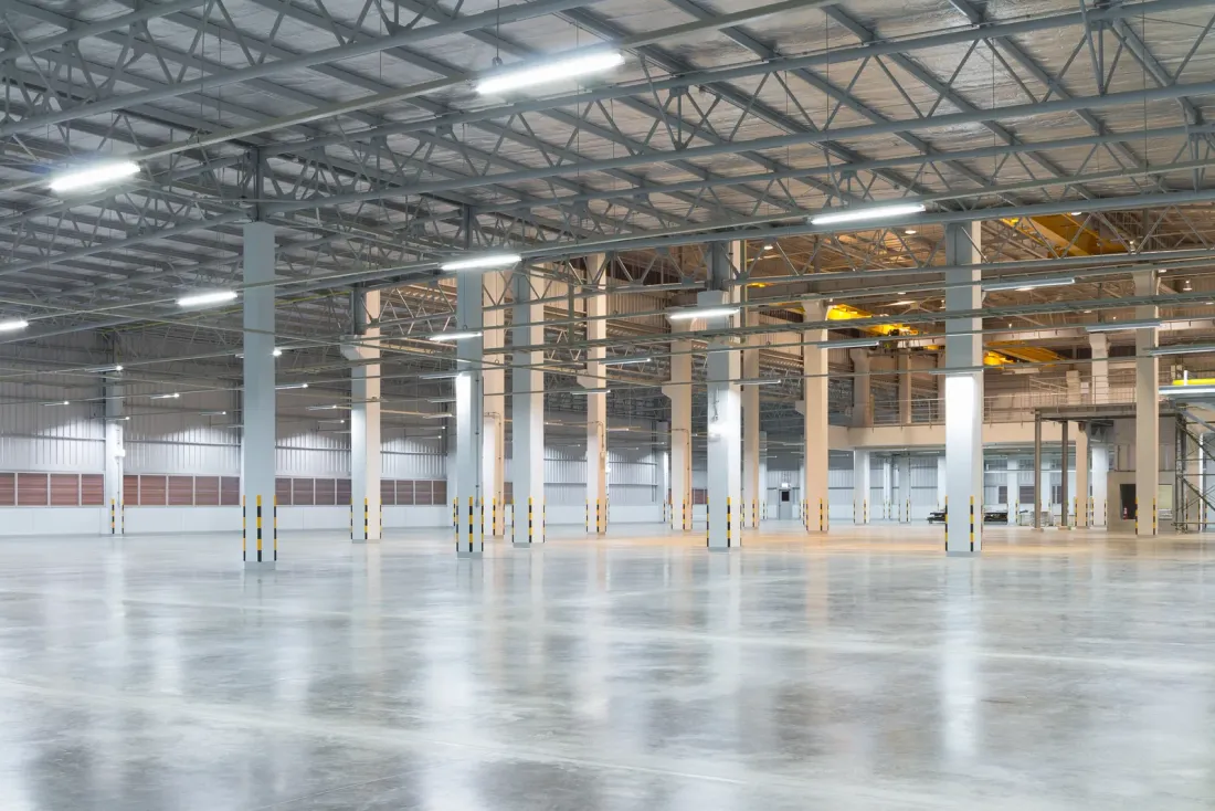 An empty warehouse with a glossy concrete floor, white columns and a steel roof structure lined with lights, a setting for commercial cleaning services in Coachella, CA