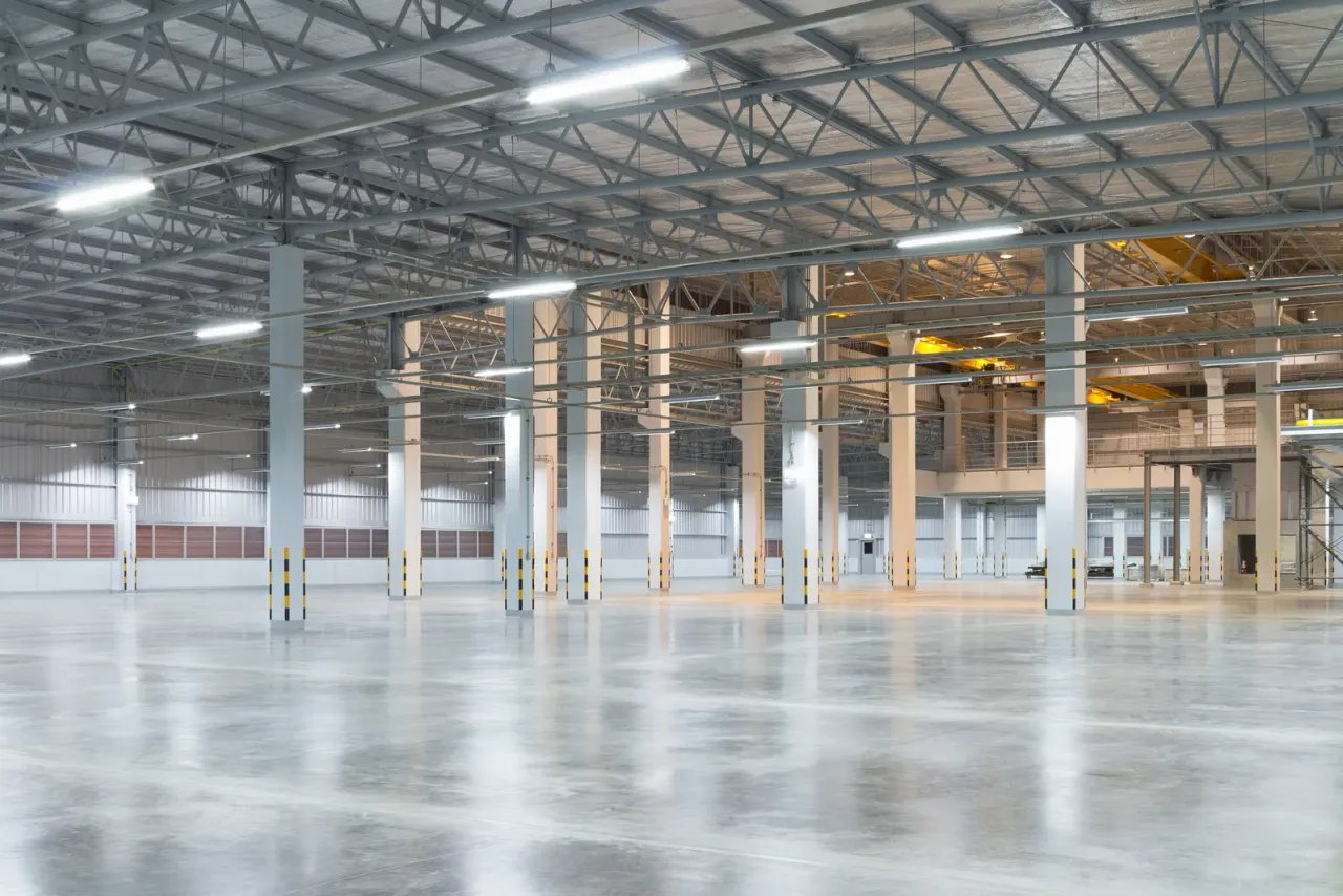 An empty warehouse with a glossy concrete floor, white columns and a steel roof structure lined with lights, the type of facility covered by warehouse cleaning in Carson, CA