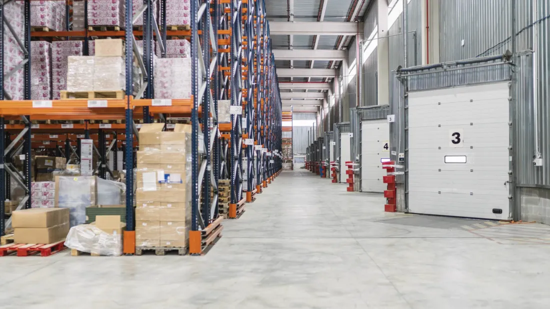 A warehouse with pallet racking stacked with boxed goods on one side and numbered loading dock doors on the other, over a clean concrete floor