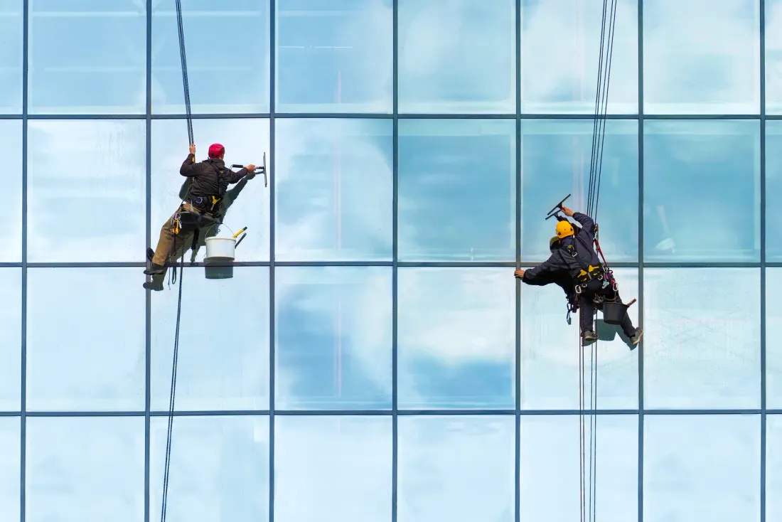 Two rope-access workers cleaning a blue glass curtain wall that reflects clouds, for a page on office cleaning services in Studio City, CA