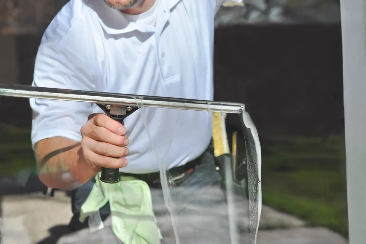 A man in a white polo shirt drawing a squeegee across a wet pane of glass, with a green cloth in his other hand, accompanying "Exterior Window Cleaning for Commercial Buildings"
